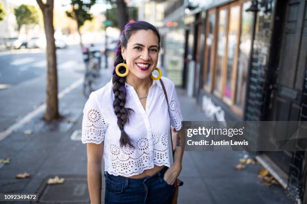 millennial latina stands alone on city sidewalk, smiling and looking at camera, wearing white lace blouse and bright yellow hoop earrings - yellow blouse foto e immagini stock