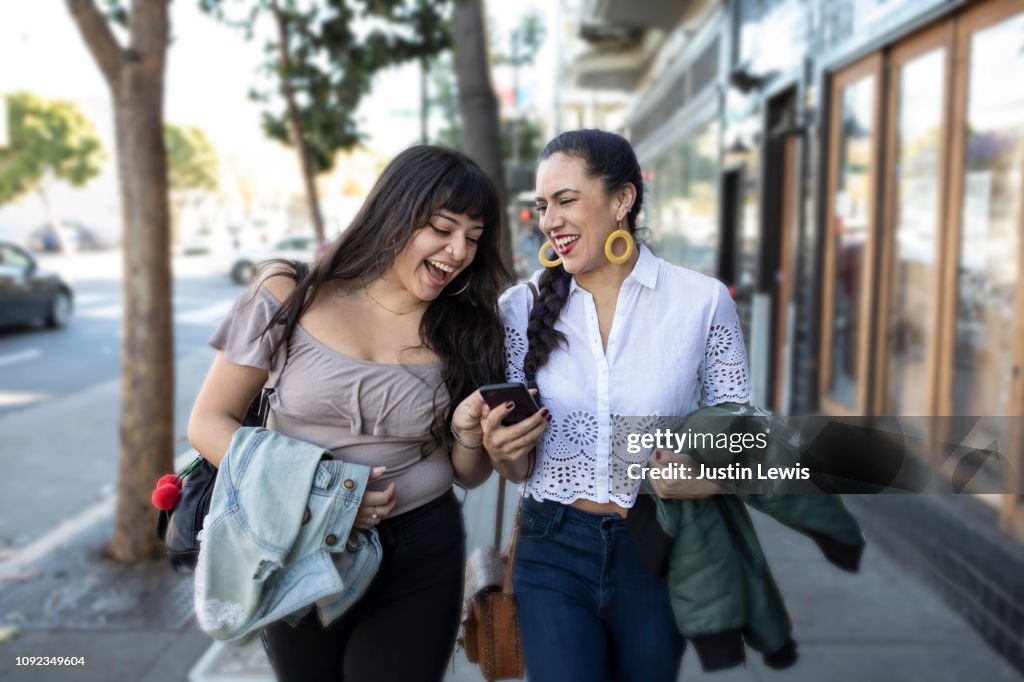 Two Latina Girlfriends Walking Along Sidewalk, Looking at Smart Phone, Laughing