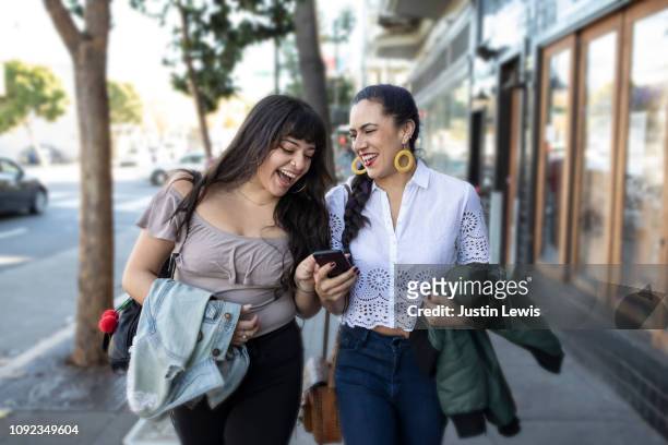 Two Latina Girlfriends Walking Along Sidewalk, Looking at Smart Phone, Laughing