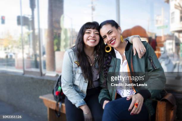 two millennial latina women lean into each other, smiling, while sitting on a bench - sister foto e immagini stock