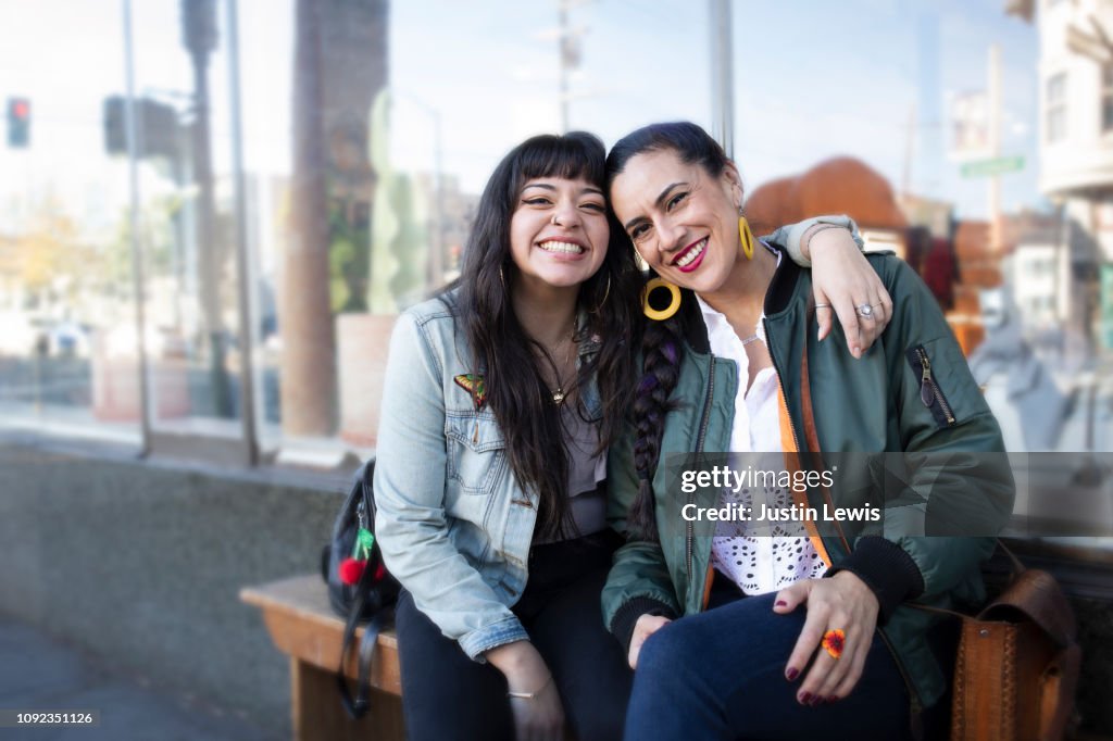 Two Millennial Latina Women Lean Into Each Other, Smiling, While Sitting on a Bench