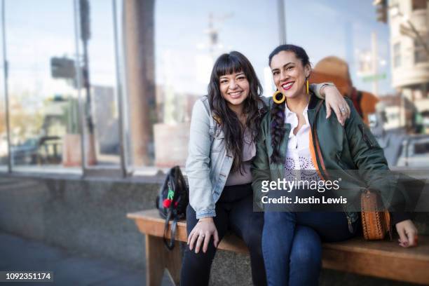 two best friends, latina millennials, sitting on a bench hugging, smiling at camera - portrait team natürlich stock-fotos und bilder
