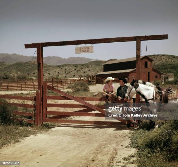 Couple, dressed in western clothing, leans against the entrance gate of the T-Bar ranch, 1950. The man holds the reins for a white horse.