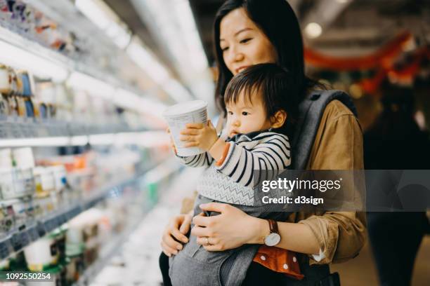 cute baby girl holding a tub of fresh yoghurt while grocery shopping with mother in a supermarket - yoghurt tub stock pictures, royalty-free photos & images