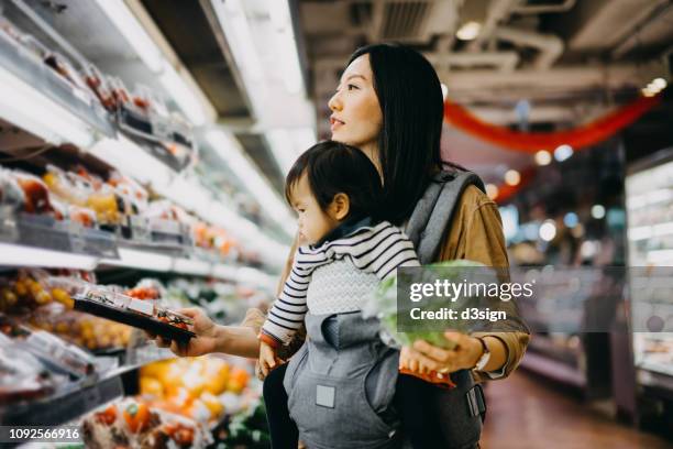 young mother grocery shopping with baby girl in supermarket, shopping for fresh organic vegetables and fruits - convenience stock pictures, royalty-free photos & images