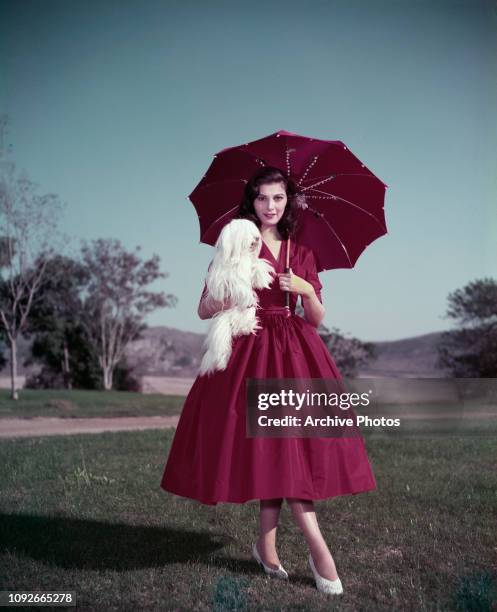 Italian actress Pier Angeli wearing a red dress and matching parasol, and holding a small white dog, circa 1955.