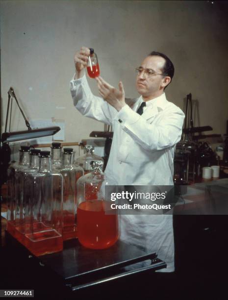 View of American scientist and physician Jonas Salk , developer of the polio vaccine, wearing a white lab coat, and smiling while holding up a bottle...