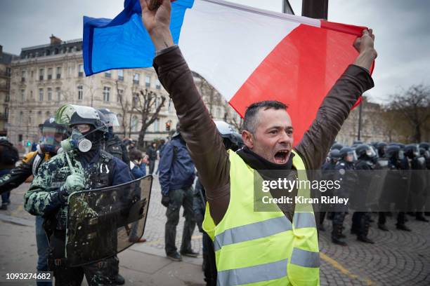 Defiant 'Gilet Jaune' or 'Yellow Vest' protestor waves the French Tricolour in front of a line of French Riot Police in another day of violence and...