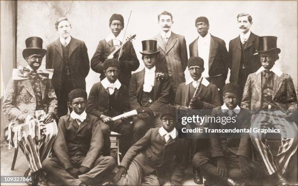 Circa 1910, Ten young men in costume and blackface, members of a minstrel troupe, pose with three others in street clothes, for a photo postcard from...