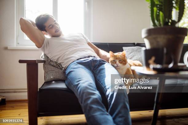 man sitting on sofa caressing his pet cat - white jeans stock pictures, royalty-free photos & images