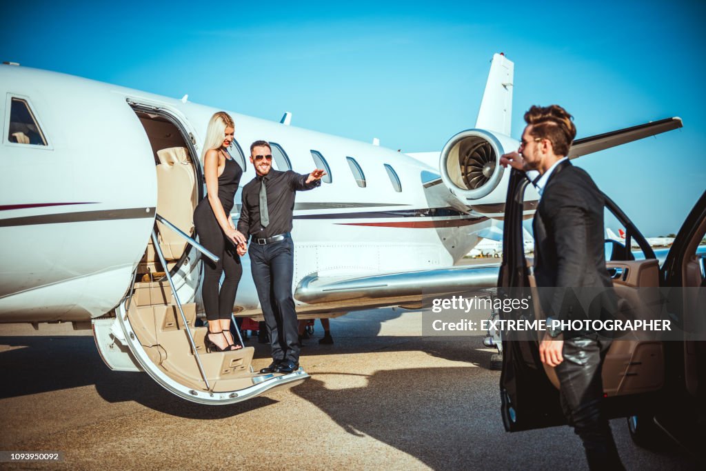 Rich business couple disembarking a private airplane while a chauffeur is waiting for them next to a car parked on an airport taxiway