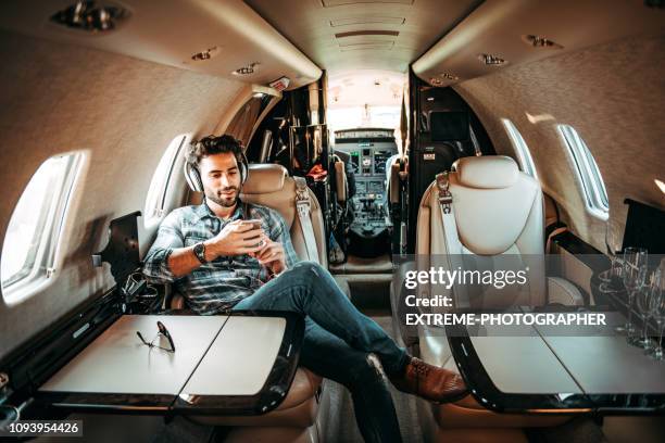 joven rico hombre escuchando música por los auriculares y el uso de un teléfono móvil mientras estaba sentado en un avión privado - avión privado fotografías e imágenes de stock