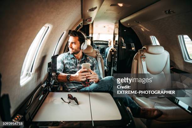 young rich man looking out of the window while listening to music over the headphones and using a smart phone, sitting in a private jet - millionnaire stock pictures, royalty-free photos & images