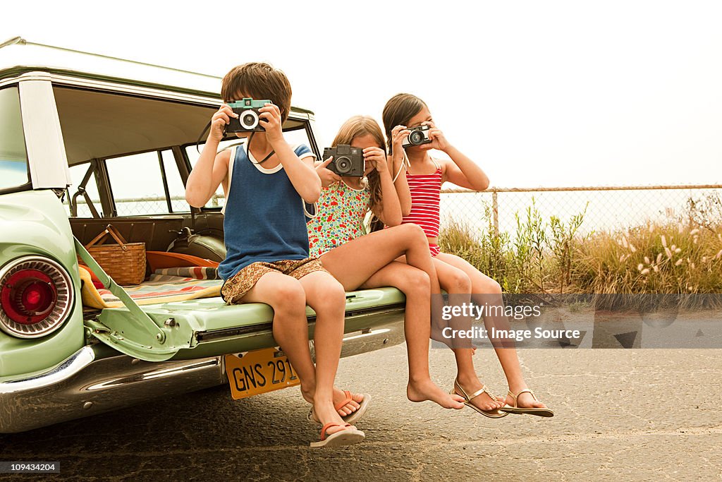 Three children sitting on back of estate car taking photographs