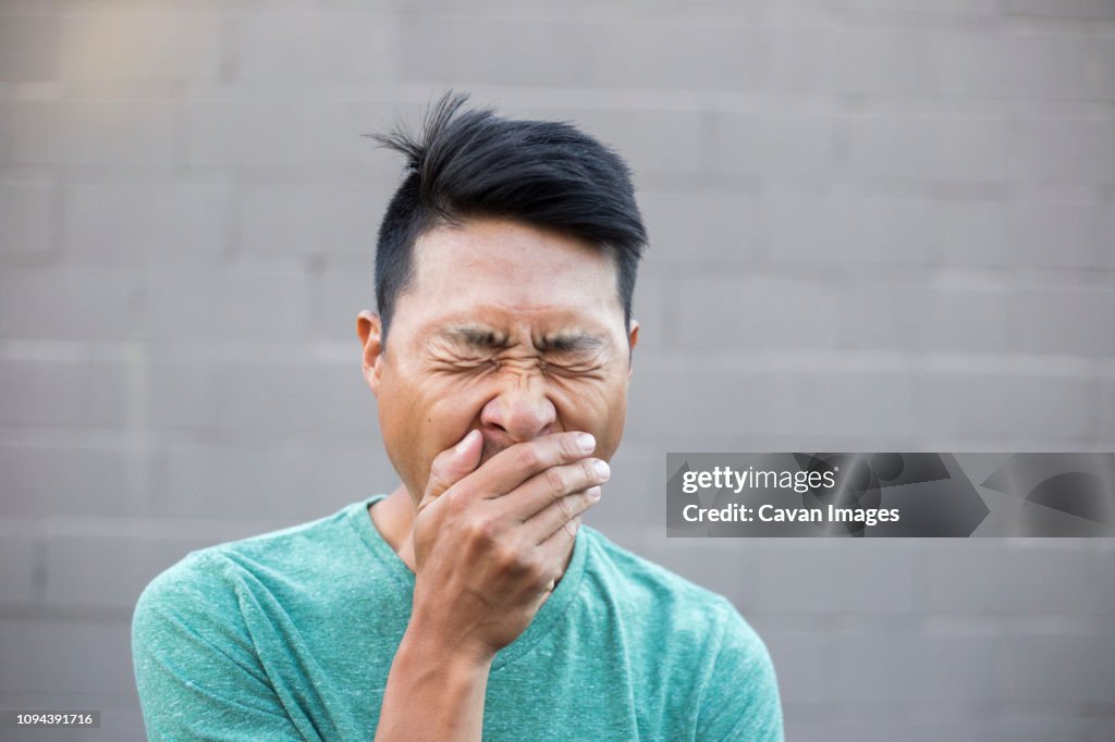 Close-up of man with hands covering mouth while yawning against wall