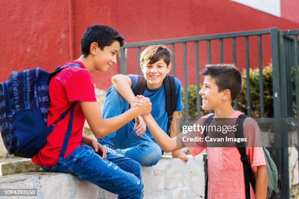 smiling boy looking at friends giving handshake while sitting on retaining wall against school building - students shaking hands stock-fotos und bilder