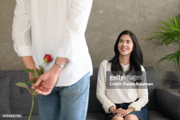 the young man is hiding flowers behind their backs to his girlfriend at home - woman giving flowers stock-fotos und bilder