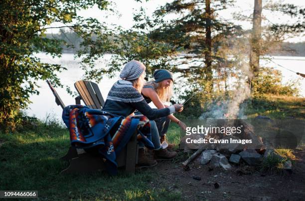 female friends cooking marshmallows over campfire while sitting on chairs in forest - ontario canada stock-fotos und bilder