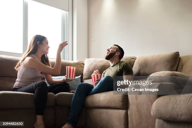 playful girlfriend throwing popcorn in boyfriend's mouth while sitting on sofa in rental house - panama food stock pictures, royalty-free photos & images