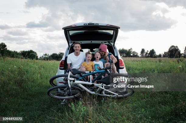 happy family with bicycles sitting on car trunk amidst grassy field against cloudy sky - auto kofferraum stock-fotos und bilder