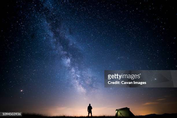 rear view of silhouette man standing on field against star field at campsite - zelt nacht stock-fotos und bilder