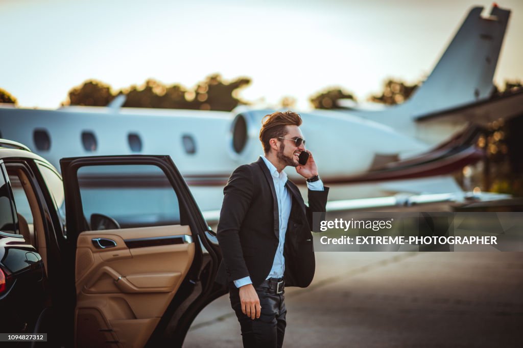 Jonge rijke zakenman praten op een mobiele telefoon tijdens het uitstappen van een luxe auto geparkeerd naast een privé vliegtuig op een asfalt