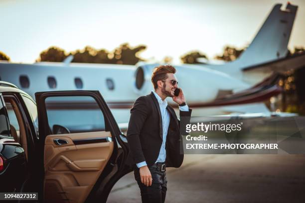 young rich businessman talking on a mobile phone while getting out of a luxurious car parked next to a private airplane on a tarmac - upper class stock pictures, royalty-free photos & images