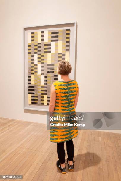 Woman wearing a brightly coloured and patterned dress looks at a piece by the weaving artist Annie Albers at a retrospective show of her work at Tate...