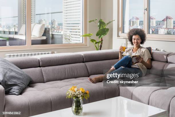 happy black woman reading a book while relaxing on sofa at home. - women of penthouse stock pictures, royalty-free photos & images