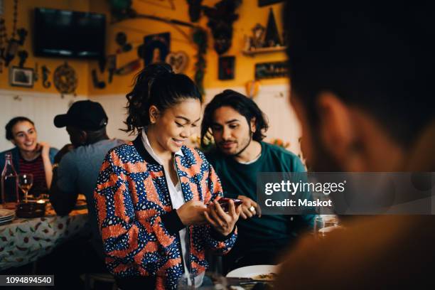 smiling young woman sharing smart phone with male friend while sitting at restaurant during dinner party - dinner at restaurant stock-fotos und bilder