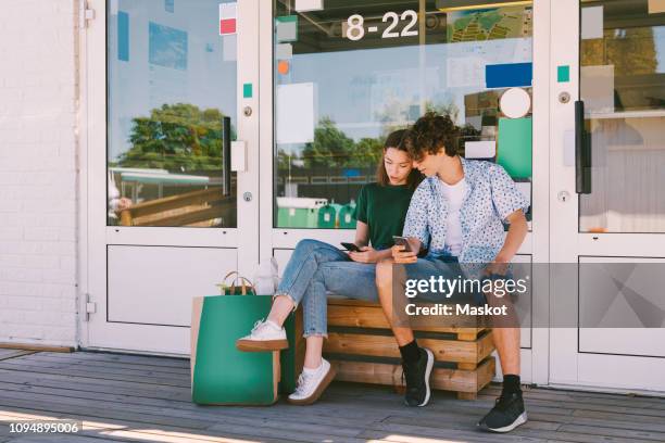 siblings using mobile phone while sitting on bench with bags outside information booth - girl who stands stock pictures, royalty-free photos & images