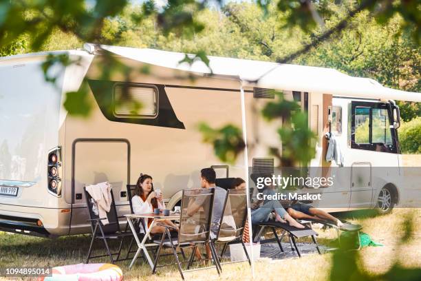 family relaxing on chairs outside camper van at campsite during summer vacation - caravan stockfoto's en -beelden