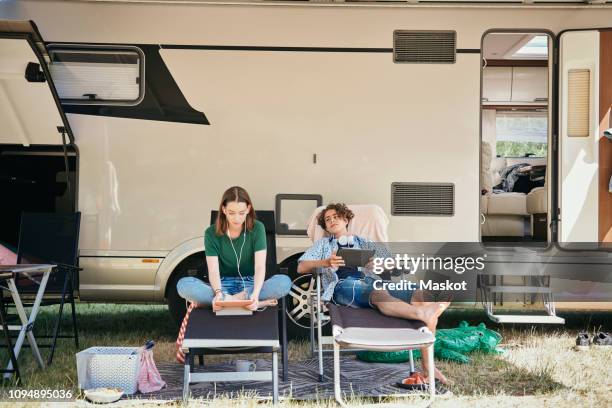 teenage siblings watching movie on digital tablet while sitting on folding chairs against motor home at campsite - mobile home photos et images de collection