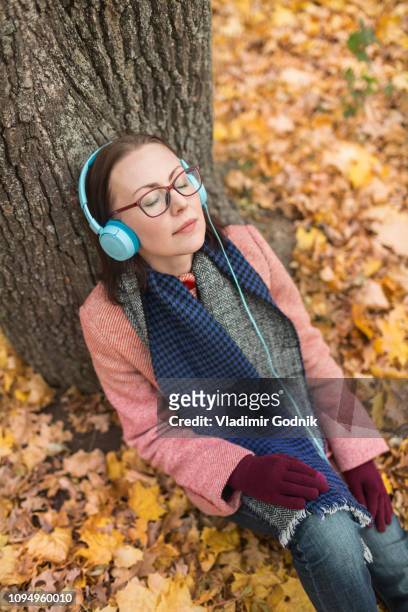 serene woman listening to music with headphones below tree in autumn park - voronezh stock pictures, royalty-free photos & images