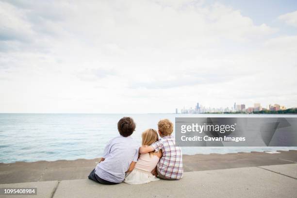 rear view of siblings sitting on promenade at beach against sky - illinois family stock pictures, royalty-free photos & images