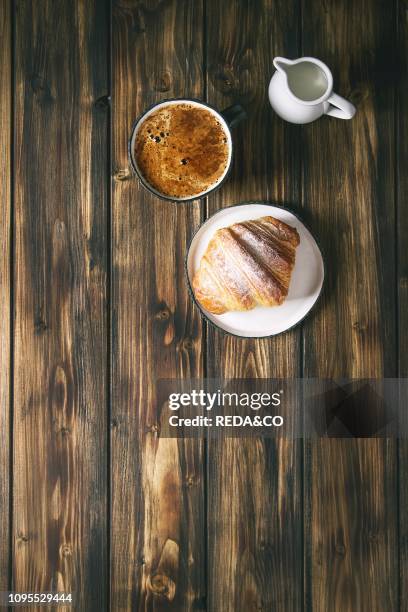 Homemade croissant with sugar powder, cup of coffee, jug of milk over wooden plank background Flat lay, space.