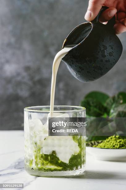 Cream pouring from jug to matcha green tea iced latte or cocktail in glass, with ice cubes, matcha powder on white marble table, decorated by green...