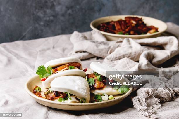 Asian sandwich steamed gua bao buns with pork belly, greens and vegetables served in ceramic plate on table with linen tablecloth Asian style fast...
