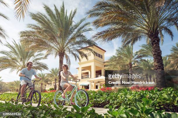 couple riding bicycles through a palm-lined luxury resort - curaçao stock pictures, royalty-free photos & images