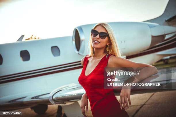 beautiful rich blonde woman leaning on a wing of a private airplane that is parked on a tarmac - millionnaire stock pictures, royalty-free photos & images