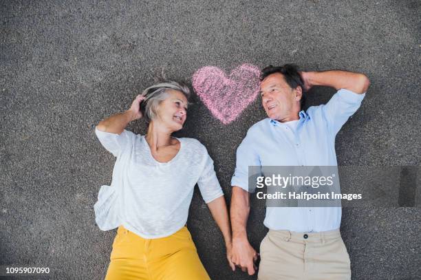 a view from directly above of senior couple lying on a road, with chalk heart overhead. - chalk heart stock pictures, royalty-free photos & images