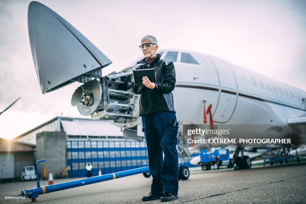 Senior female engineer holding digital tablet next to a a small private airplane with an opened nose cone parked on a taxiway