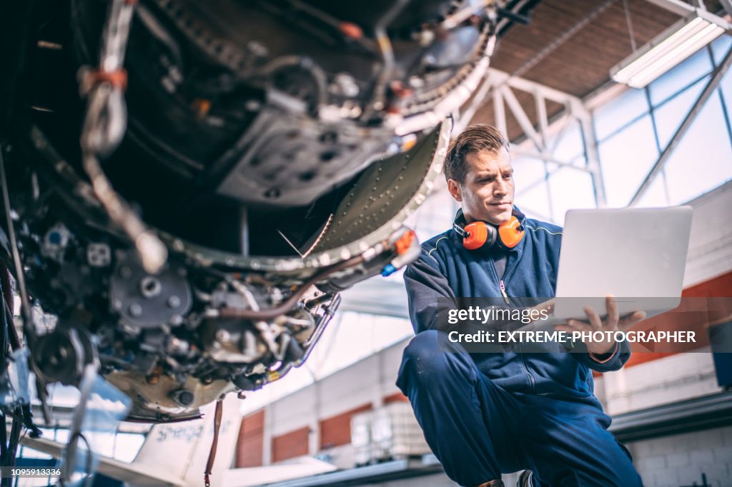 Aircraft mechanic doing some diagnostics on a laptop next to a jet engine in an aircraft hangar