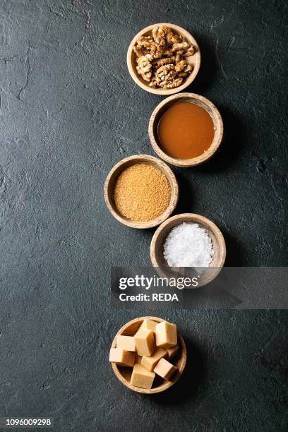 Salted caramel fudge candy served with fleur de sel, caramel sauce, brown cane sugar and caramelized walnuts in wood bowls over black texture...