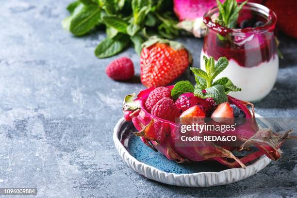 Vegan fruit salad with berries and mint served in pink dragon fruit with ingredients above on ceramic plate over blue texture background Close up...