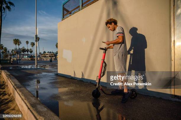 The young Latino man uses his smartphone to book electrical scooter at Venice Beach, California, USA