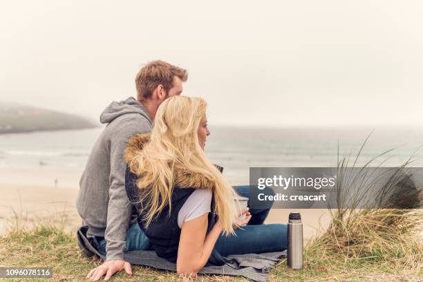 happy couple sat on sand dunes above fistral beach, newquay enjoying a cup of coffee from a flask on an autumn afternoon. - couple dunes stock pictures, royalty-free photos & images