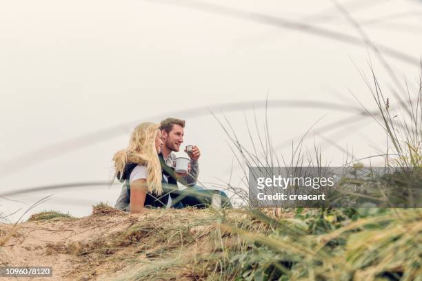 glückliches paar saß auf sanddünen über fistral beach, newquay, genießen eine tasse kaffee aus einer flasche an einem herbst-nachmittag. - couple dunes stock-fotos und bilder