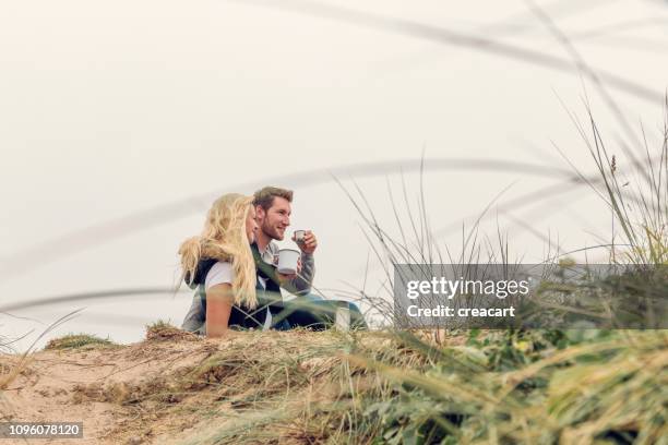 happy couple sat on sand dunes above fistral beach, newquay enjoying a cup of coffee from a flask on an autumn afternoon. - couple dunes stock pictures, royalty-free photos & images