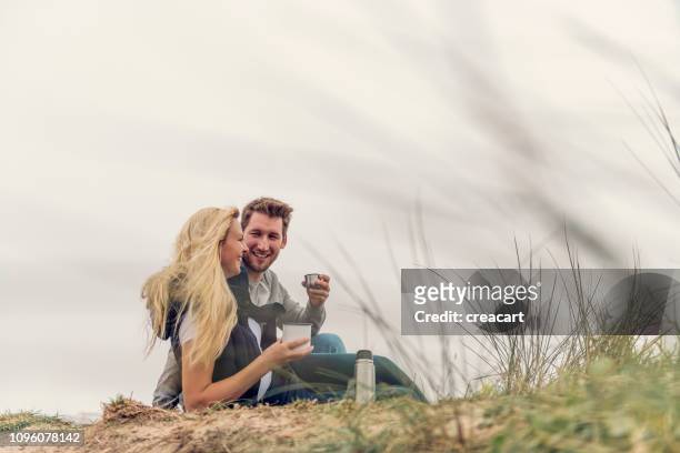 gelukkige paar zat op zandduinen boven fistral beach, newquay, genieten van een kopje koffie uit een kolf op een middag van de herfst. - couple dunes stockfoto's en -beelden